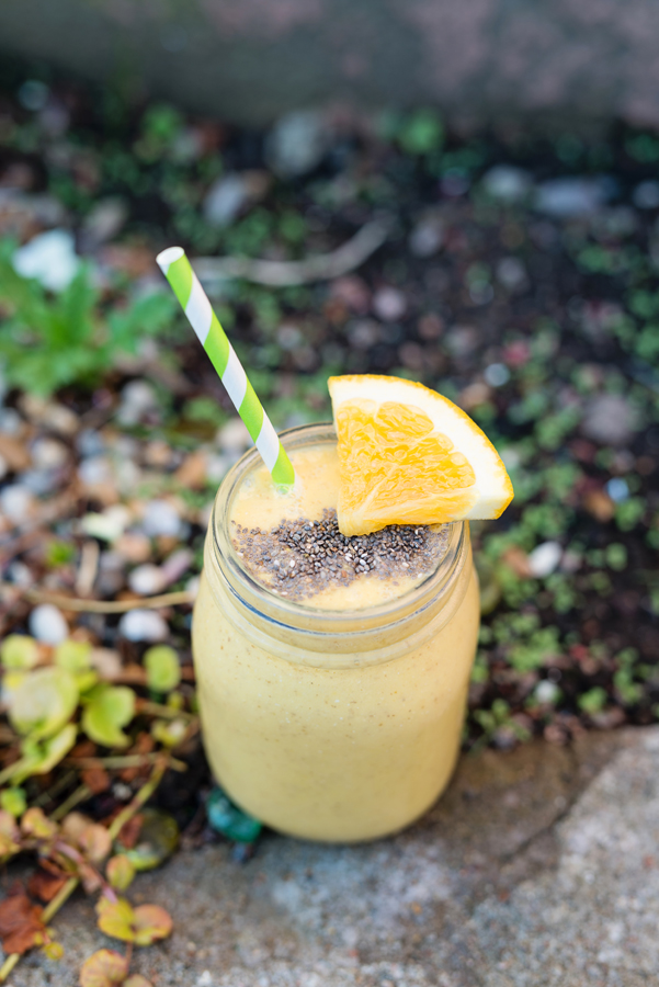 En gul smoothie med en citronskiva, chiafrön och ett pappersugrör ståendes på en sten utomhus.